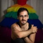 תמונת פורטרט של מתן אנחיסי, מרצה במחלקה לעיצוב אופנה. מתן נראה כאשר מאחוריו שמלה עם חצאית בצבעי הקשת