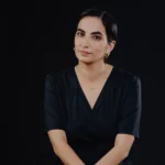 תמונת פורטרט של שקד ארקין, משמרת ומנהלת ארכיון האופנה והטקסטיל בשנקר. שקד נראית לבושה בשמלת מעטפת שחורה על רקע שחור